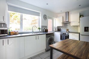 A modern kitchen with white cabinets, a built-in washing machine, a wooden dining table, and a large window providing natural light. - Fully Refurbished North Belfast Home (Belfast)