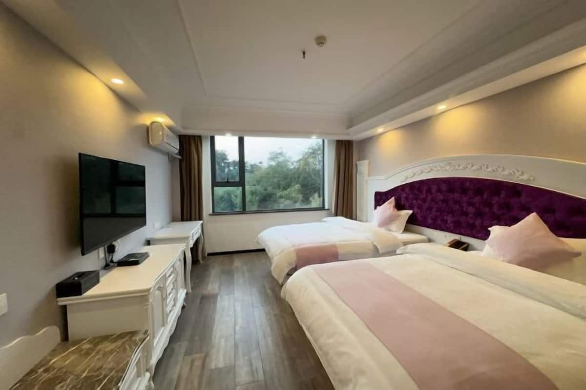 A hotel room with two beds, a TV, a desk, and a window with a view of trees.