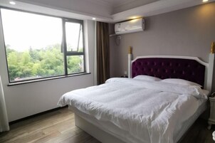 Quarto com uma janela grande, uma cama com roupa de cama branca, cabeceira roxa e um aparelho de ar condicionado na parede.