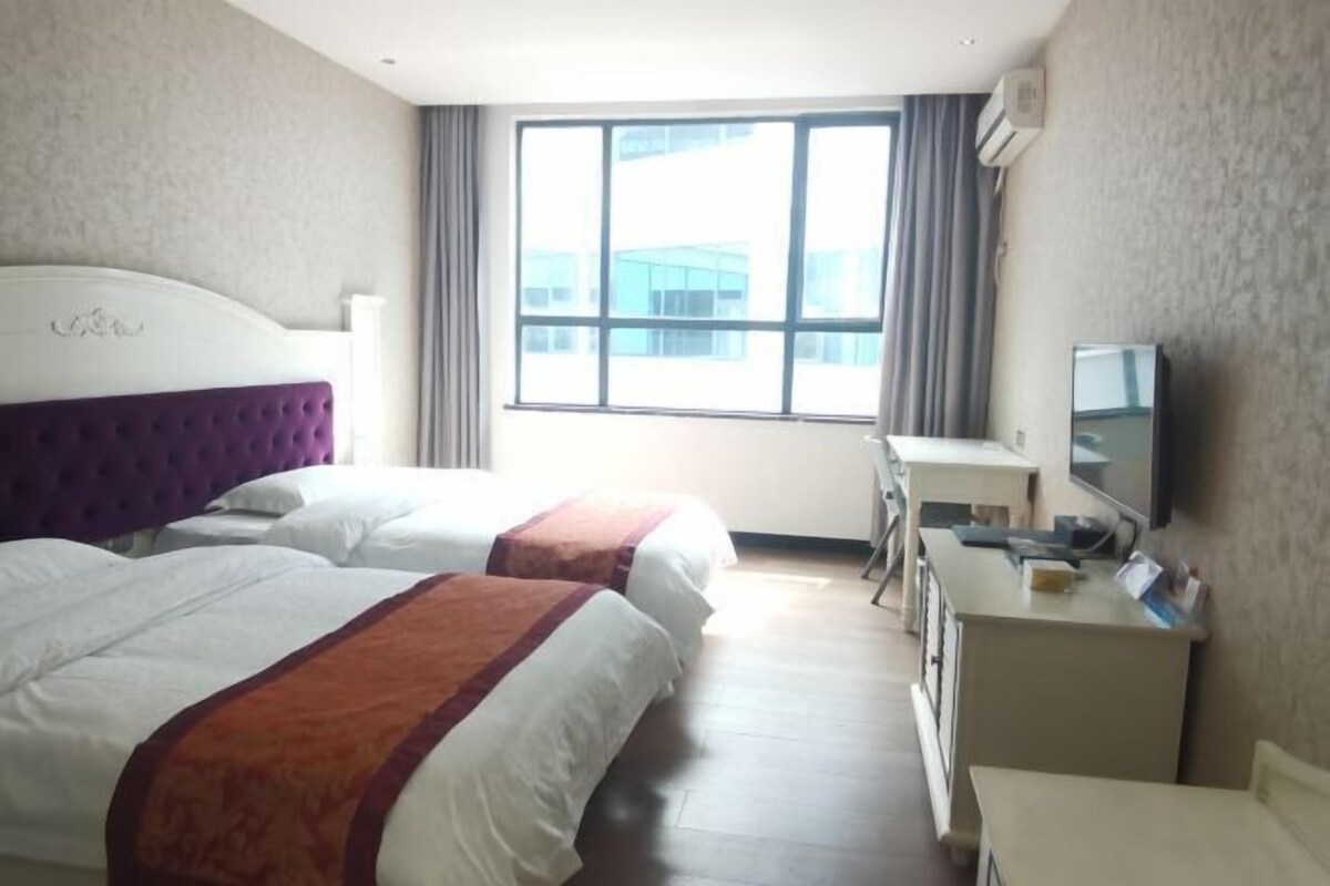 A hotel room with two beds, a desk, a television, and a large window with a view of buildings.