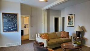 Een woonkamer met een beige bank, een salontafel en een lamp. Aan de muur hangt een prikbord met tekeningen en tekst.