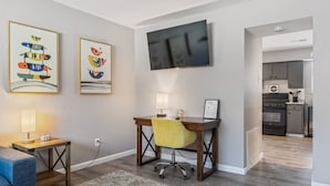A modern living room with a wooden desk, a yellow chair, a TV mounted on the wall, and a kitchen visible through an open doorway.