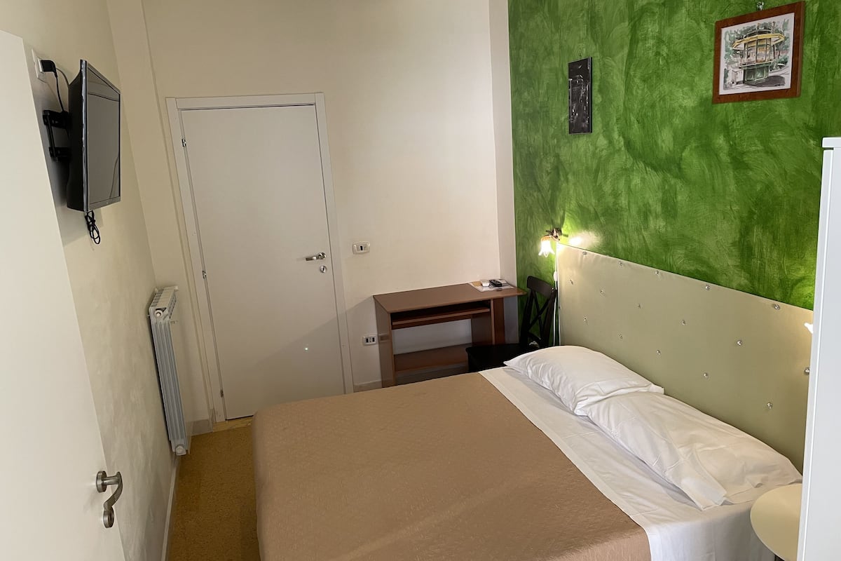 Hotelkamer met een bed, bureau, stoel, radiator, televisie en een groene muur met een afbeelding.