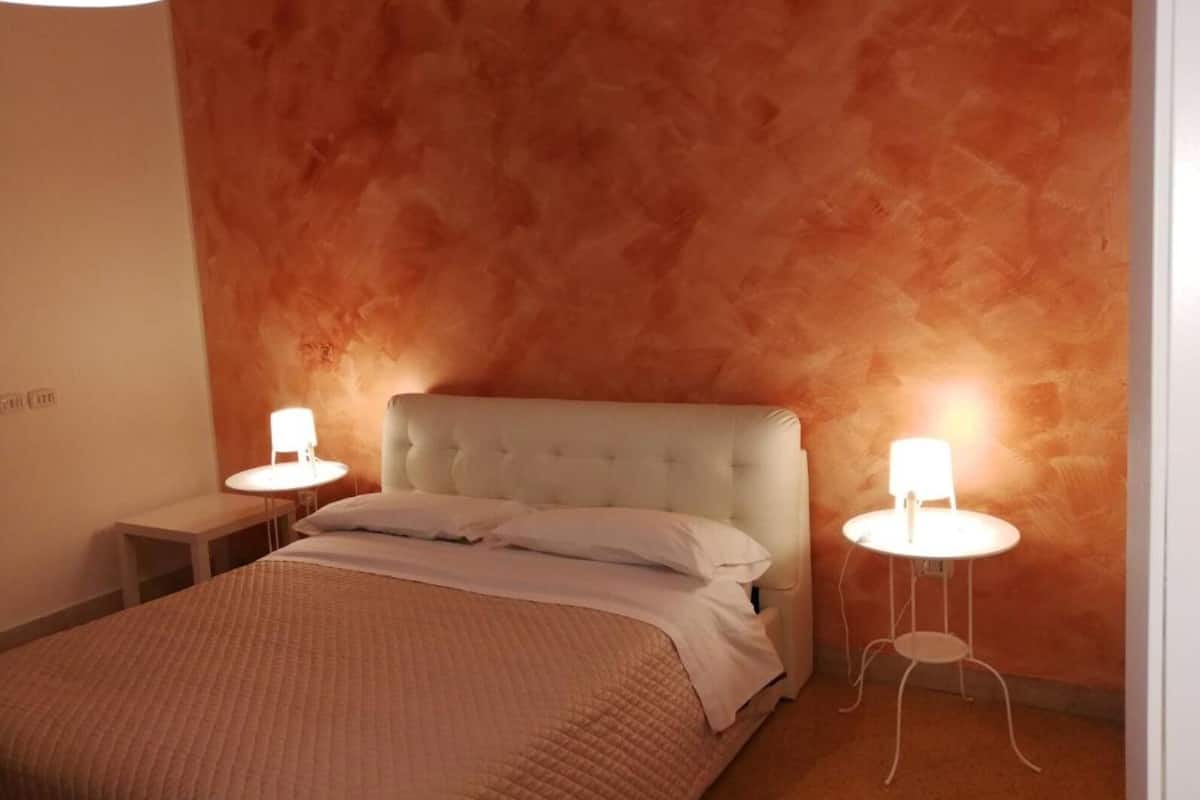Een slaapkamer met een gestoffeerd hoofdeinde, een bed met een gewatteerde deken en twee nachtkastjes met lampen.