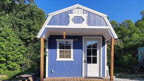 A small, single-story building with a blue exterior and white trim. It features a front door, a small window, and a porch area with a wooden bench and a lattice fence. - Unique, Cozy Stay near Red River Gorge (Slade)