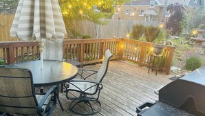 Eine Holzterrasse mit rundem Tisch und Stühlen, einem Grill und Lichterketten.