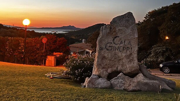 A stone monument with "Cala de Ginepri" inscribed, surrounded by grass and a sunset in the background. - Villas Smeraldas Holiday In Sardinia (Sardegna)