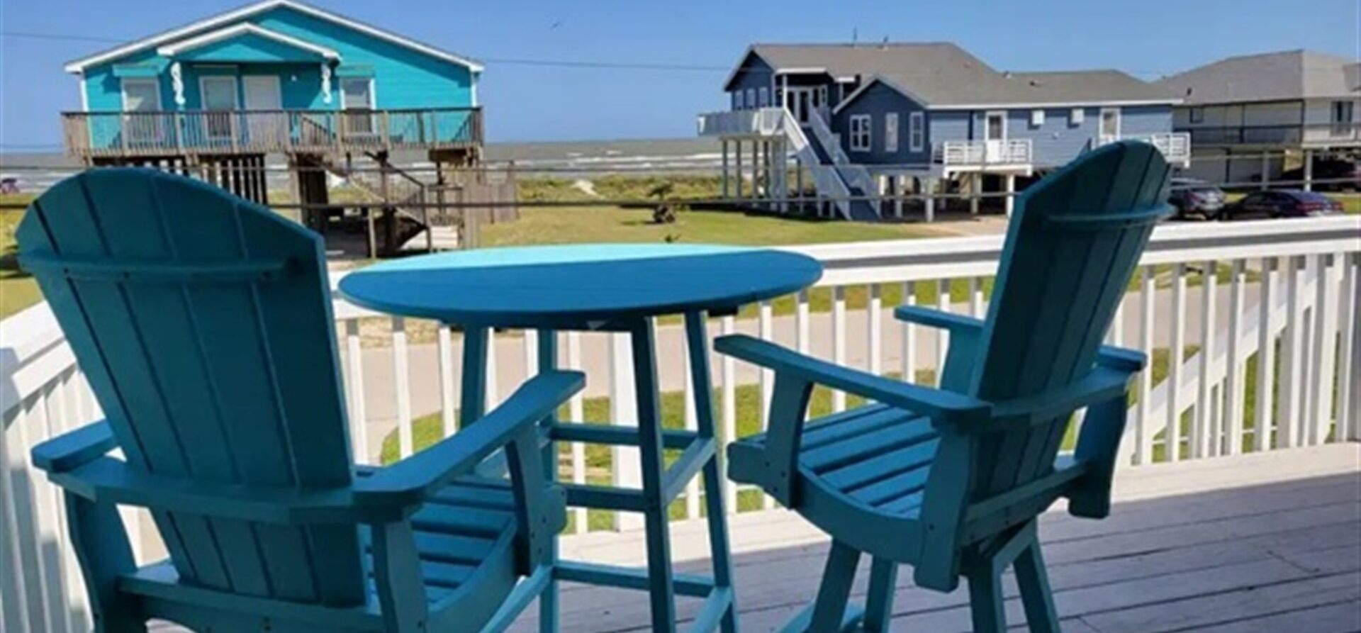 A deck with two chairs and a small table, overlooking a beach and houses.