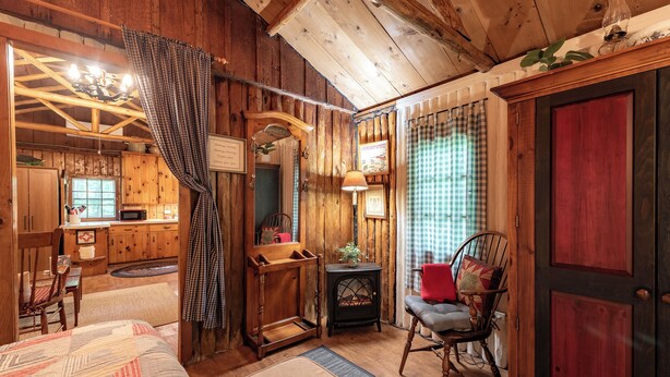 Een eenvoudig houten blokhutinterieur met houten wanden, een bed, een schommelstoel, een tafeltje en een open haard.