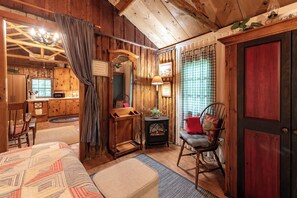 A rustic cabin interior with wooden walls, a bed, a rocking chair, a small table, and a fireplace.