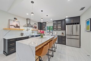 A modern kitchen with a marble island, bar stools, dark cabinetry, stainless steel appliances, pendant lighting, and artwork on the walls. - Luxury 3/3 with Heated Pool, Spa, & Putting Green!  (Sarasota)