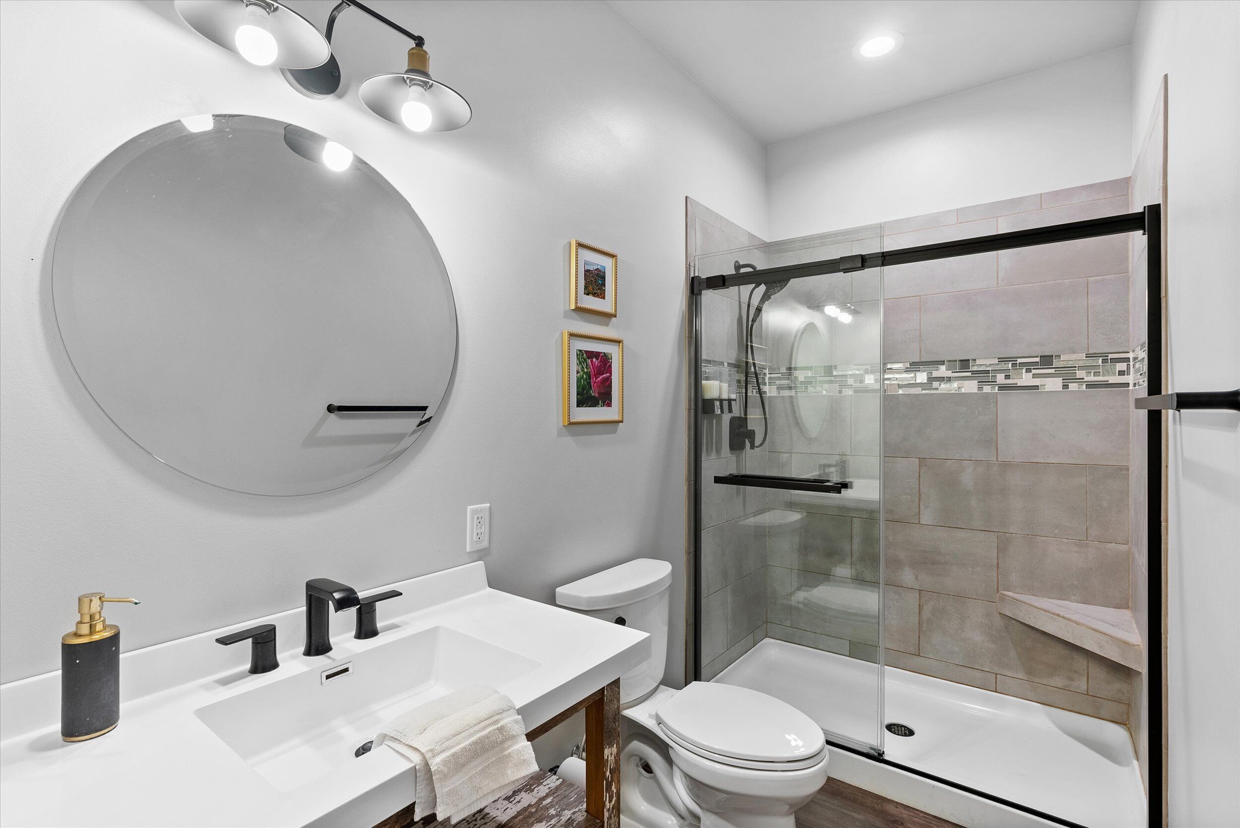 A modern bathroom with a round mirror, white sink, black faucet, and a glass-enclosed shower.