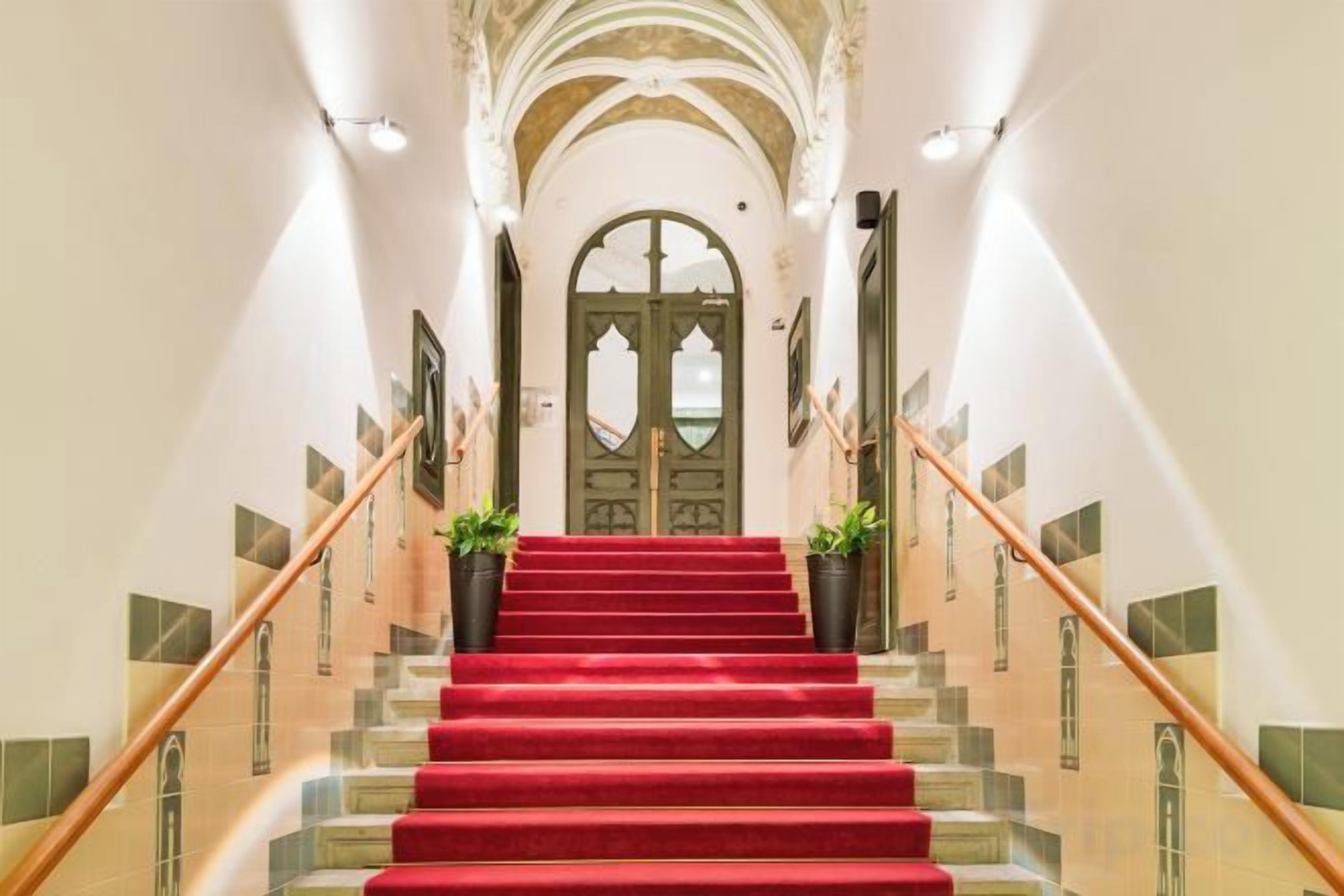Un magnifique escalier, avec des marches recouvertes de moquette rouge, menant à une grande porte en arc de cercle.