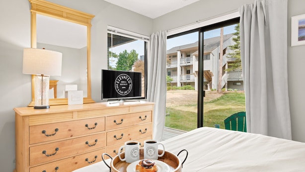 Une chambre d’hôtel avec un lit, une commode en bois avec des tiroirs, une télévision, un plateau avec deux tasses et une pâtisserie, et une grande fenêtre donnant sur des bâtiments et de la verdure.