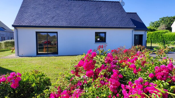 Une maison moderne avec un toit foncé, de grandes fenêtres en verre et un jardin bien entretenu agrémenté de fleurs roses éclatantes.