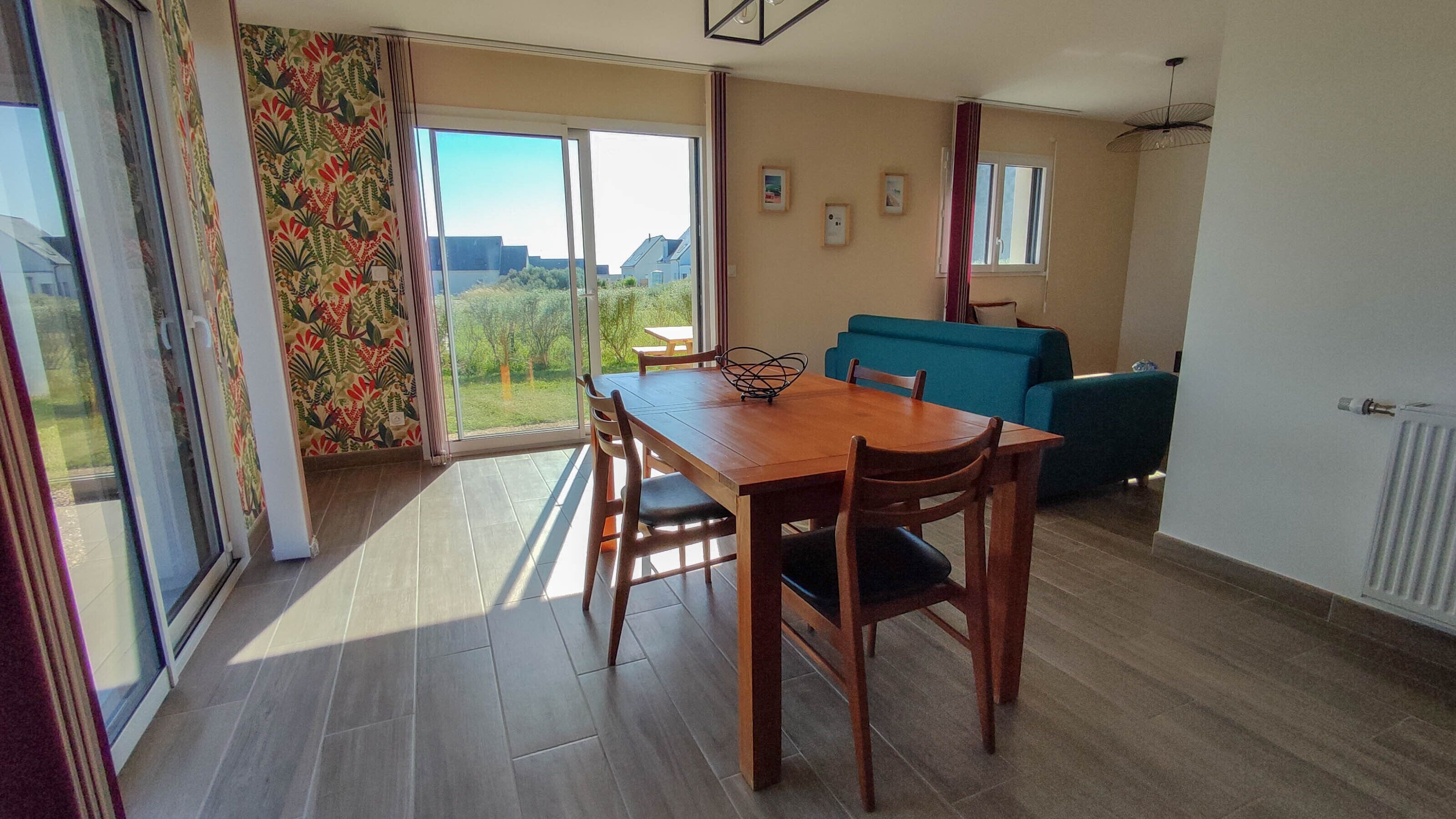 A dining area with a wooden table and chairs, a blue sofa, and a view of greenery through large windows.