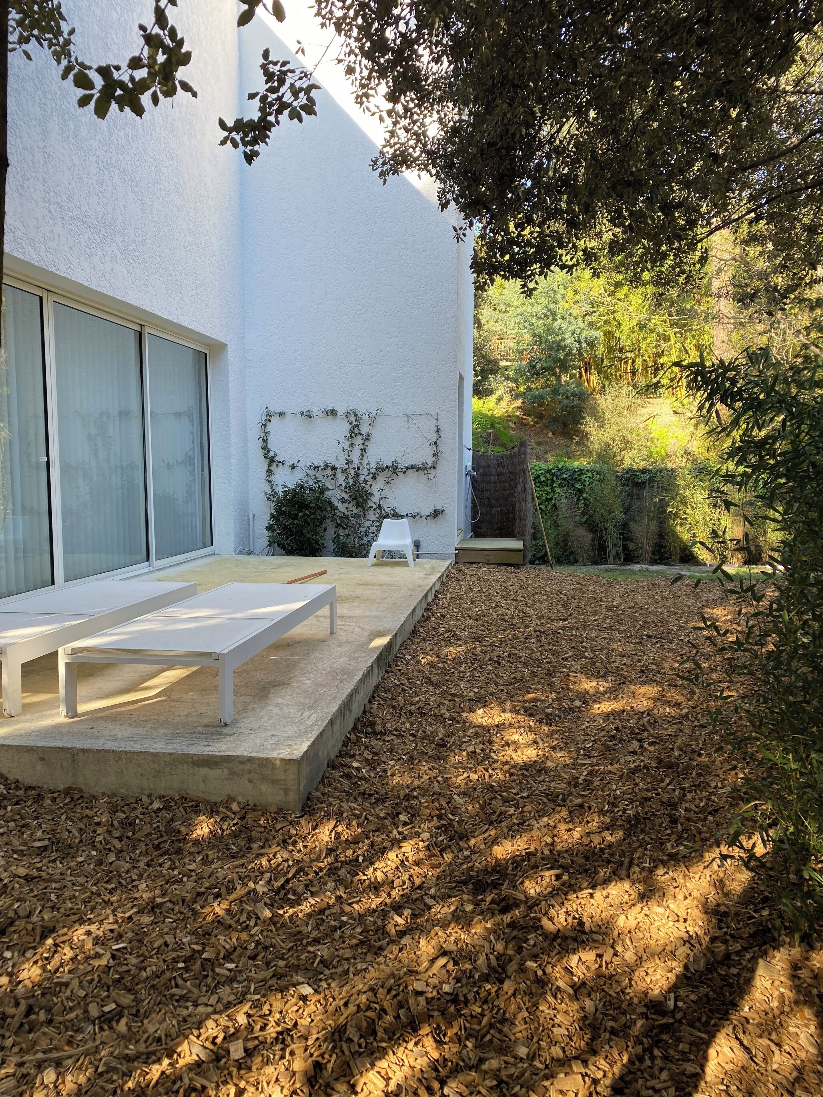 A patio area with a white bench and a table, surrounded by wood chips and greenery.