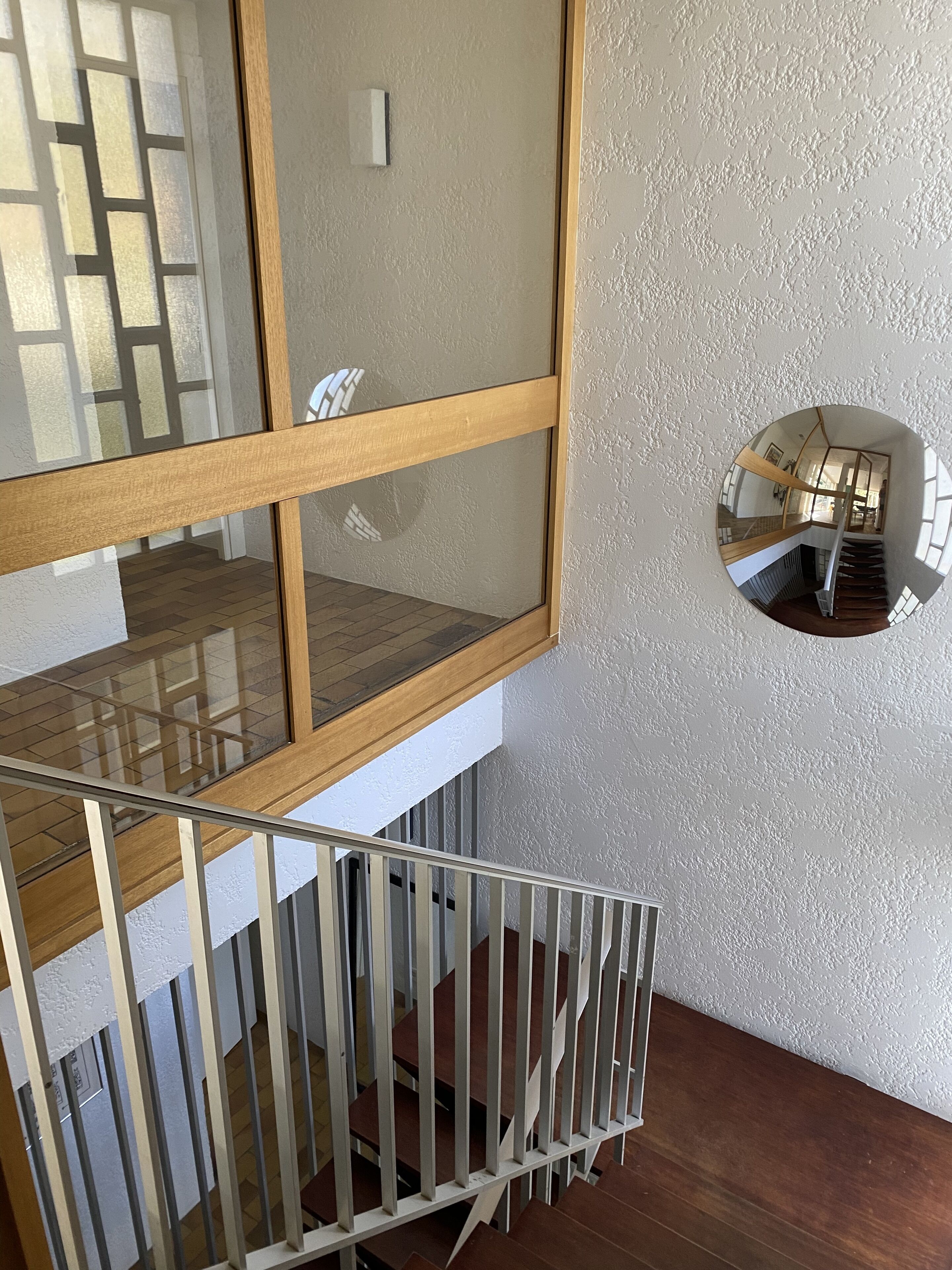Een modern interieur met een glazen en houten trap, een ronde spiegel aan de muur, en zicht op de bovenverdieping door het glas.