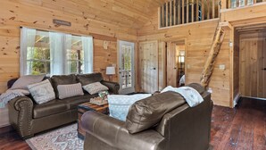Un intérieur de chalet chaleureux, avec un revêtement mural en bois, une chambre à coucher située en mezzanine accessible par un escalier en bois, et un espace de vie comprenant un canapé et une table basse.