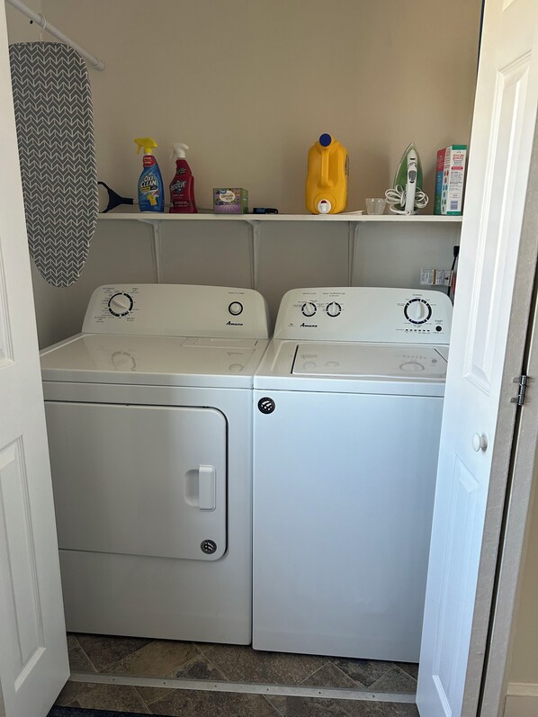 A laundry area with two front-loading washing machines, a shelf with cleaning supplies, and a patterned shower curtain. - Sailwinds Loft at Cambridge Creek (Cambridge)