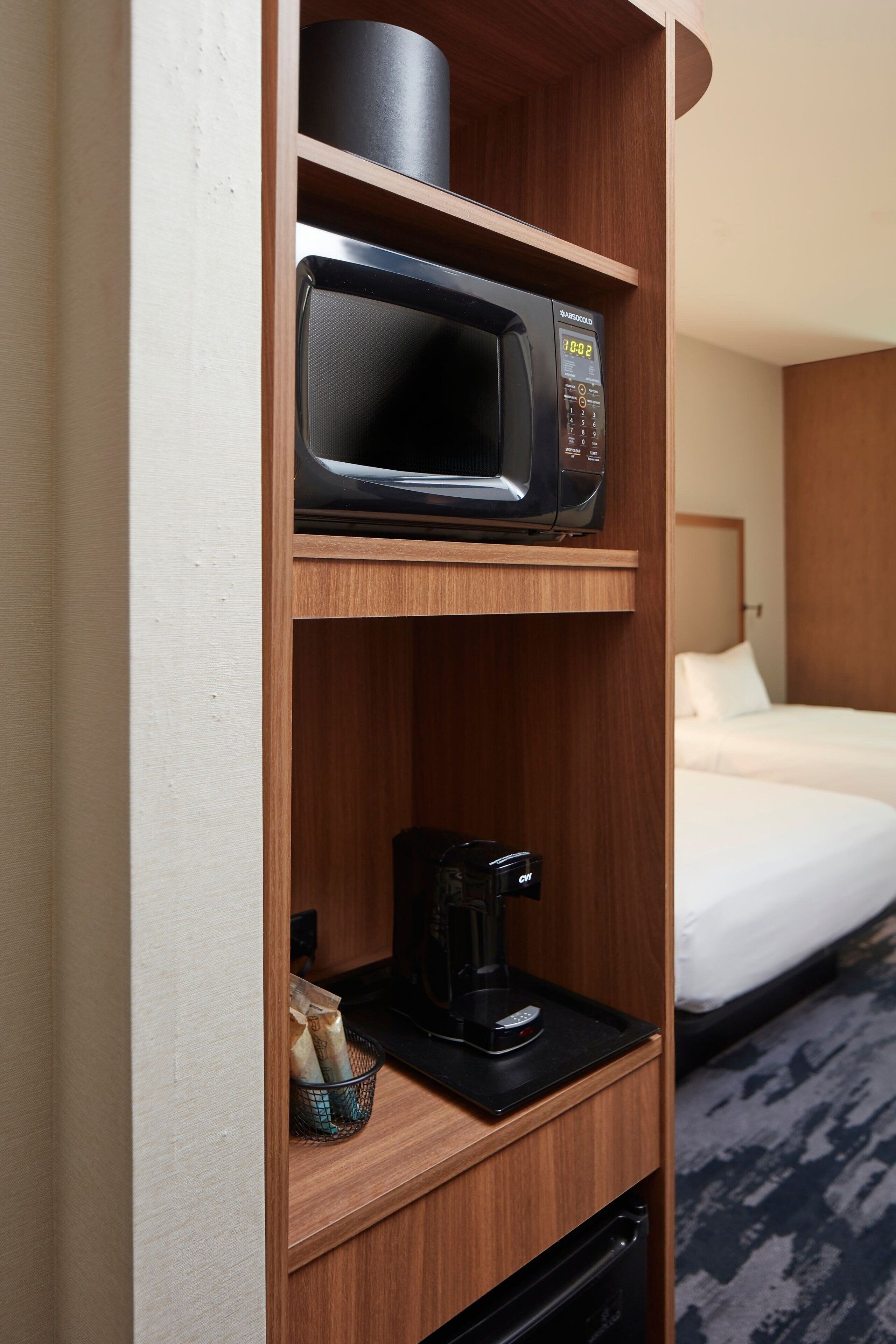 A hotel room with a microwave on a shelf, a coffee maker, and a basket with items.