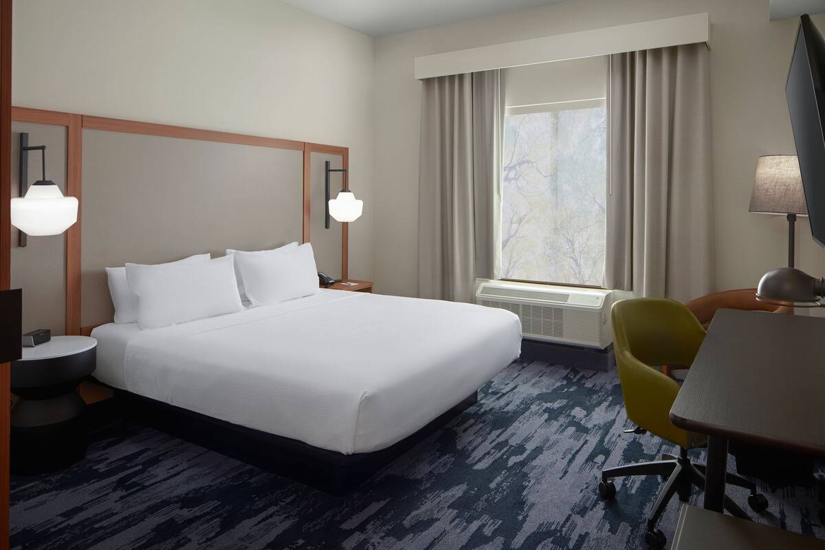 A hotel room with a large bed, a desk, a chair, a lamp, and a window with curtains.