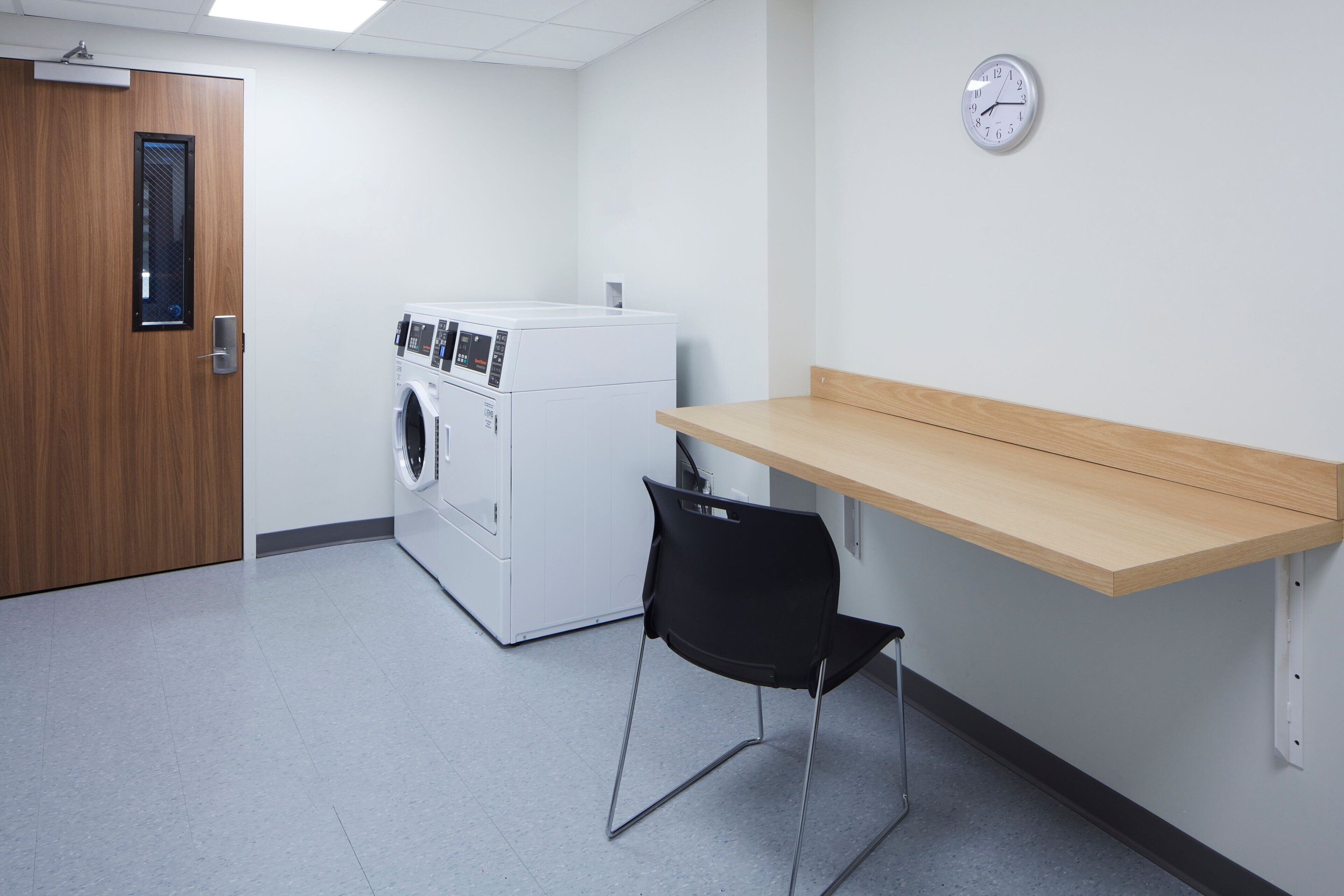 A laundry room with a washing machine, a dryer, a wooden bench, and a wall clock.