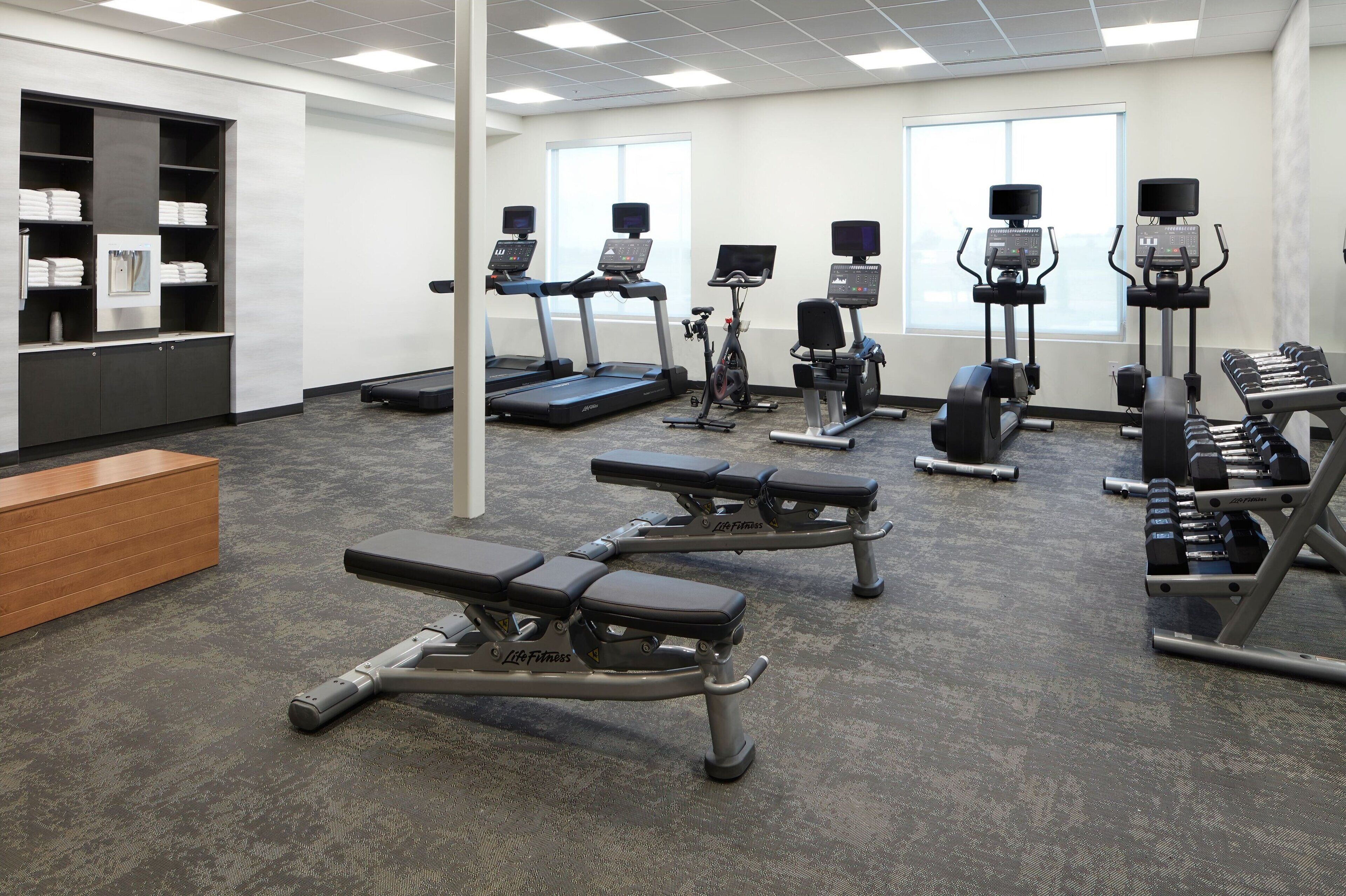 A gym with treadmills, elliptical machines, weight benches, and a wooden bench.