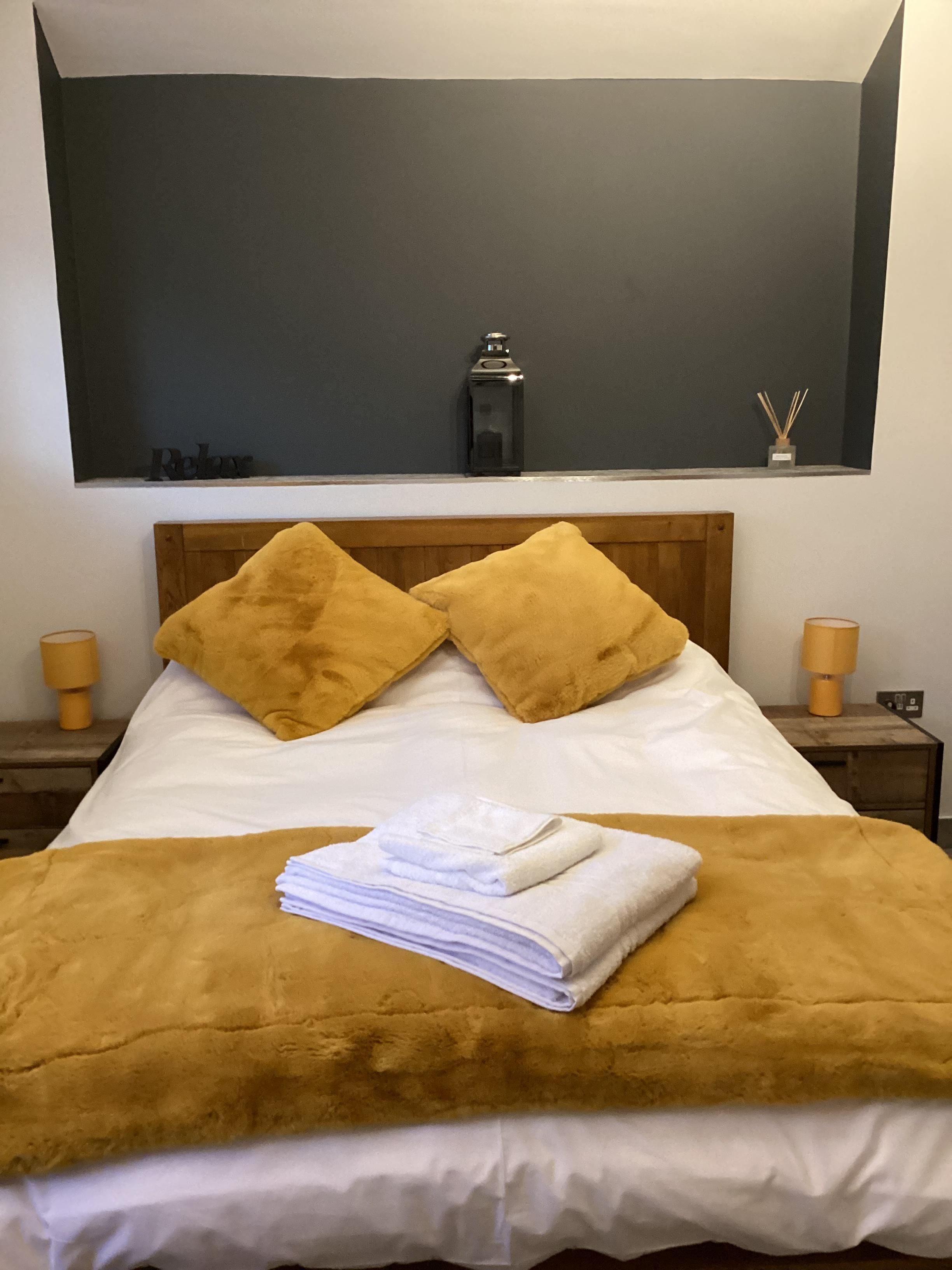Een keurig opgemaakt bed met een bruin hoofdbord, wit beddengoed en twee bruine kussens. Aan de zijkant staat een klein tafeltje met een lamp en een decoratief object.