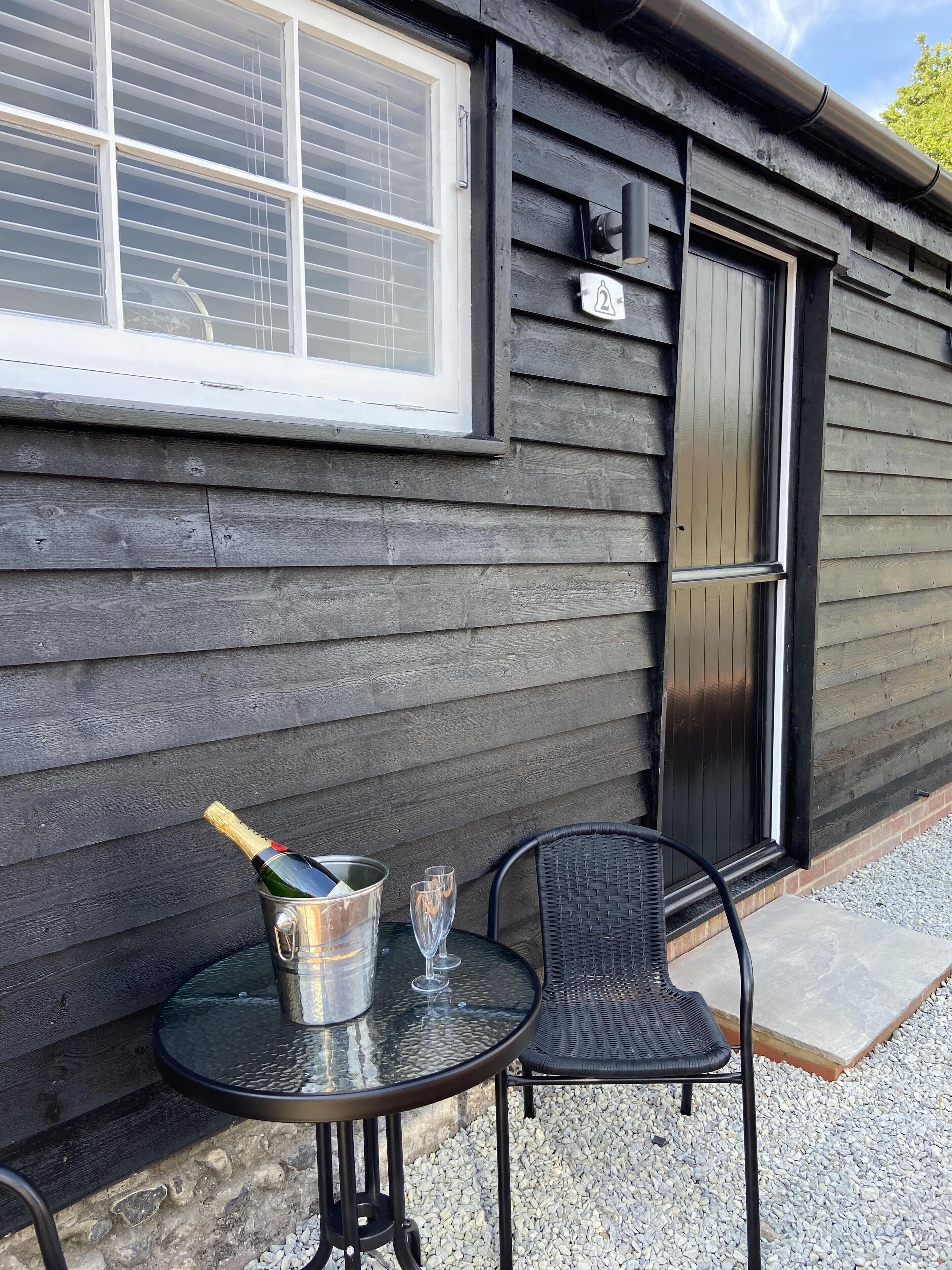 A small outdoor table with a metal bucket containing a bottle and glasses, positioned in front of a wooden building with a window and a door.