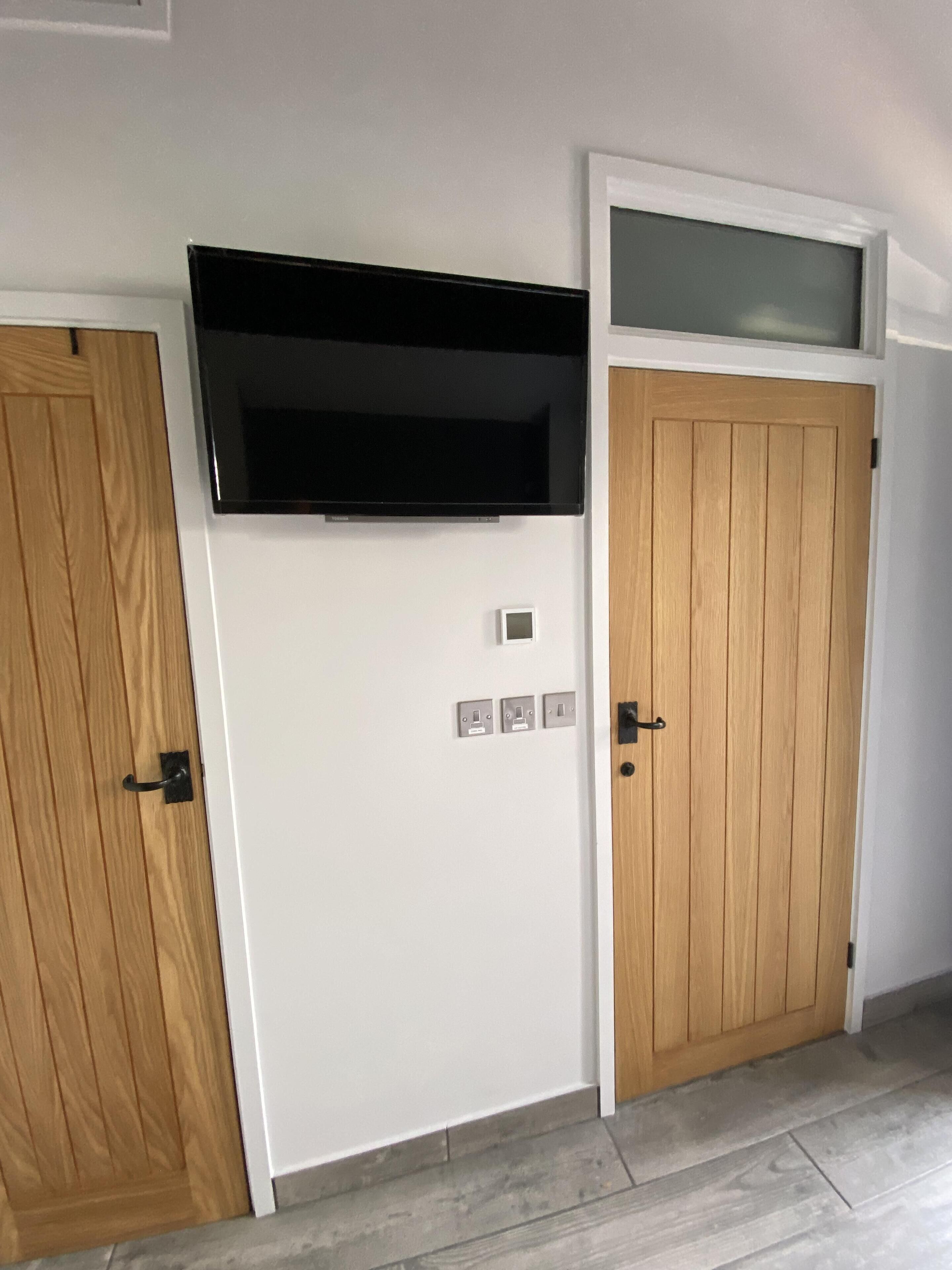 Une pièce avec un mur blanc, un téléviseur à écran plat fixé au mur et deux portes en bois, avec une petite fenêtre au-dessus de la porte de droite.