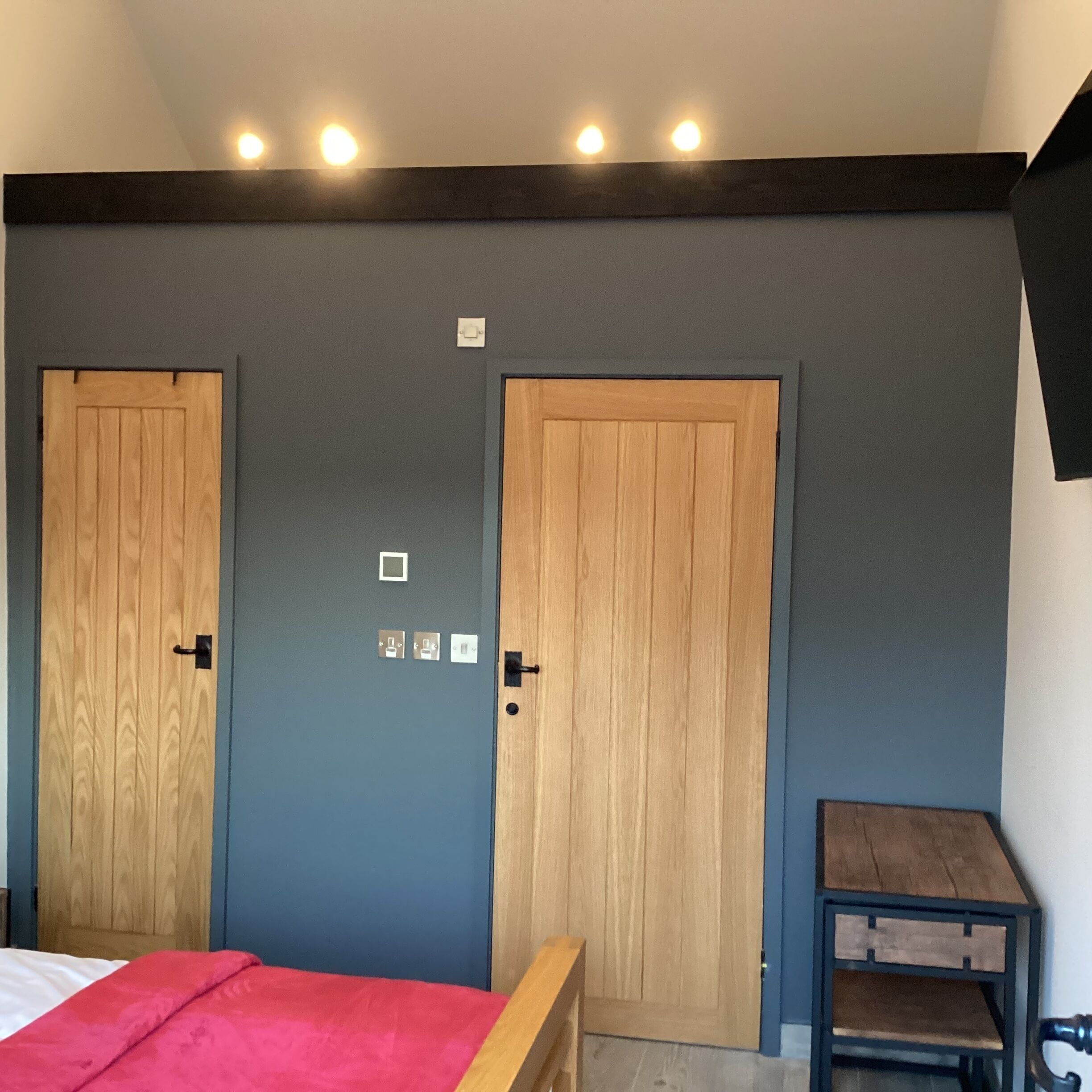 A bedroom with two wooden doors, a bed with a red blanket, a small table, and a wall-mounted light fixture.
