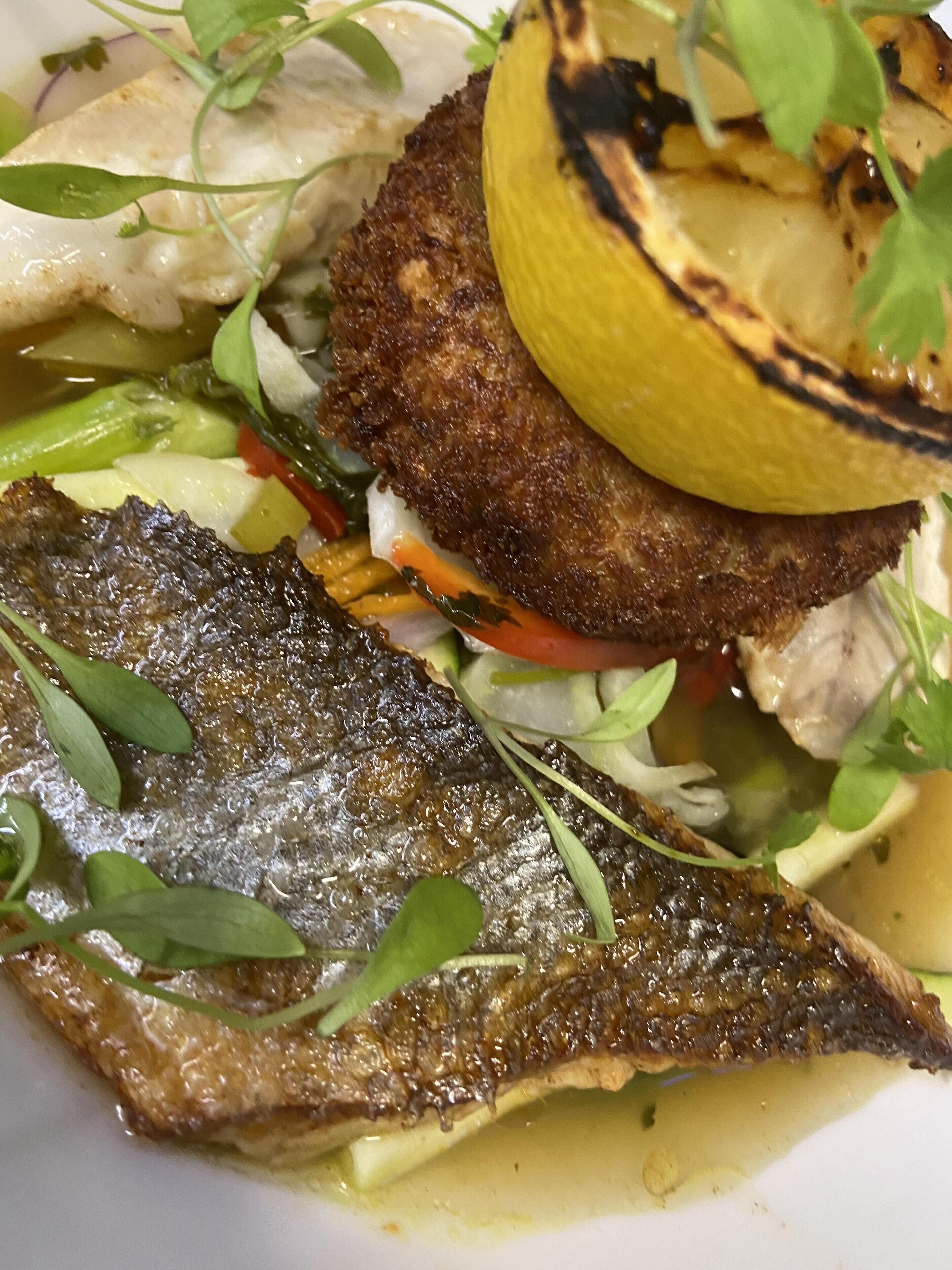 A plate of food with grilled fish, vegetables, and a slice of grilled lemon.