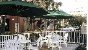 Buiten zitplaatsen met witte plastic stoelen en groene parasols, een wit hek en een bord in de verte.
