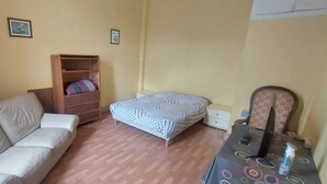 Una habitación con sofá, cama, un armario de madera, un ventilador y una mesa con una botella.