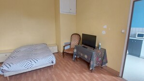 Un dormitorio con cama, una silla, una mesa con televisor y un rincón de kitchenette.