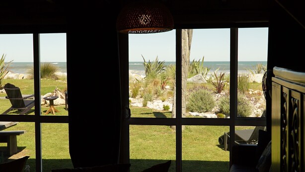 Habitación con amplias ventanas que dan a una playa, con una silla, una mesita y un ventilador.