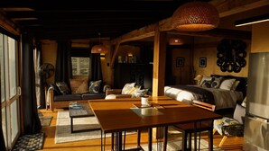 Una habitación acogedora y rústica con una mesa de comedor de madera, sofá y cama. Hay lámparas con pantalla de mimbre, un ventilador y un refrigerador.
