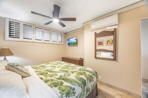 A bedroom with a bed, a ceiling fan, a TV, and a window with shutters.