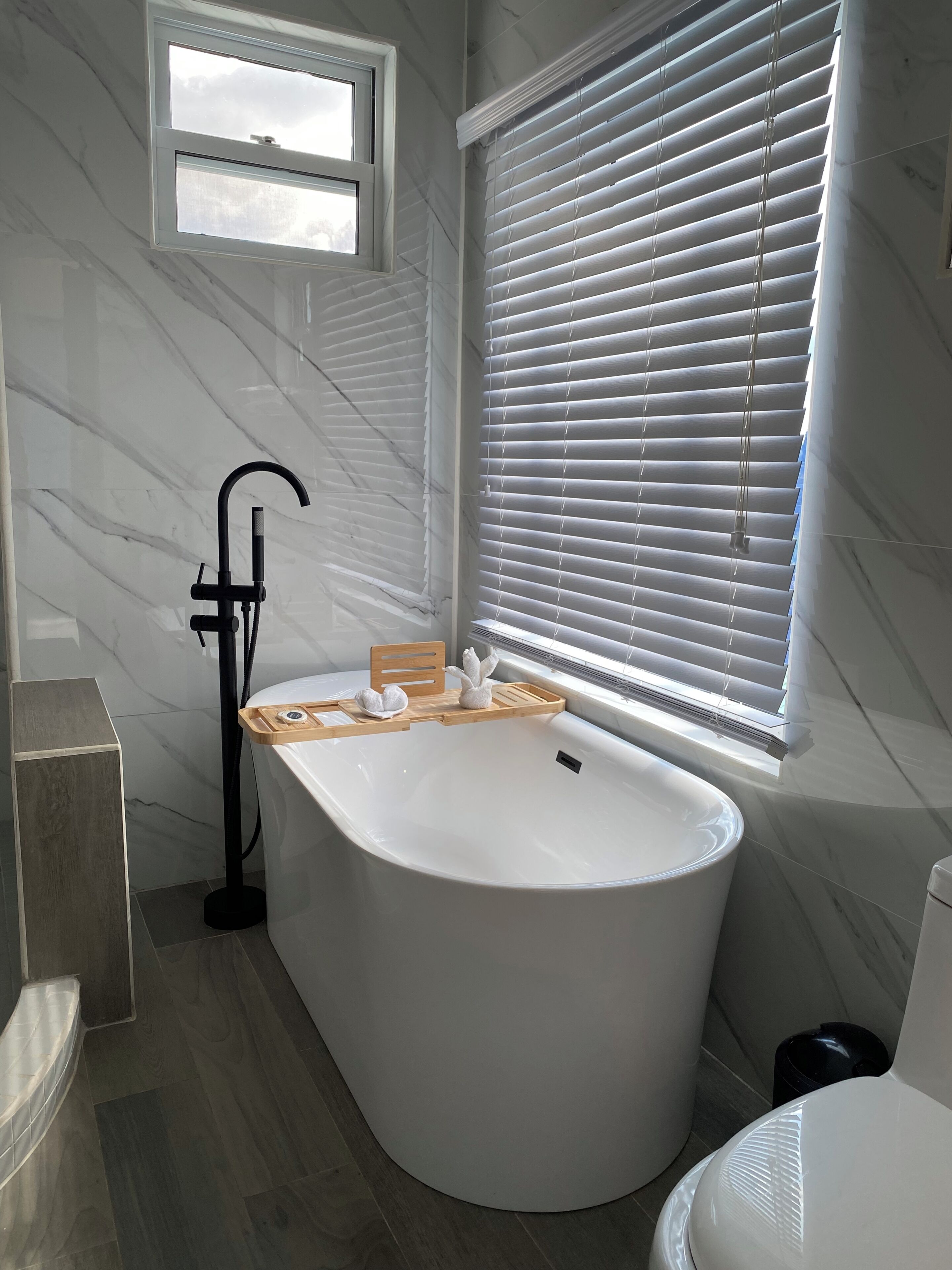 A modern bathroom with a freestanding bathtub, a window with blinds, and a marble wall.