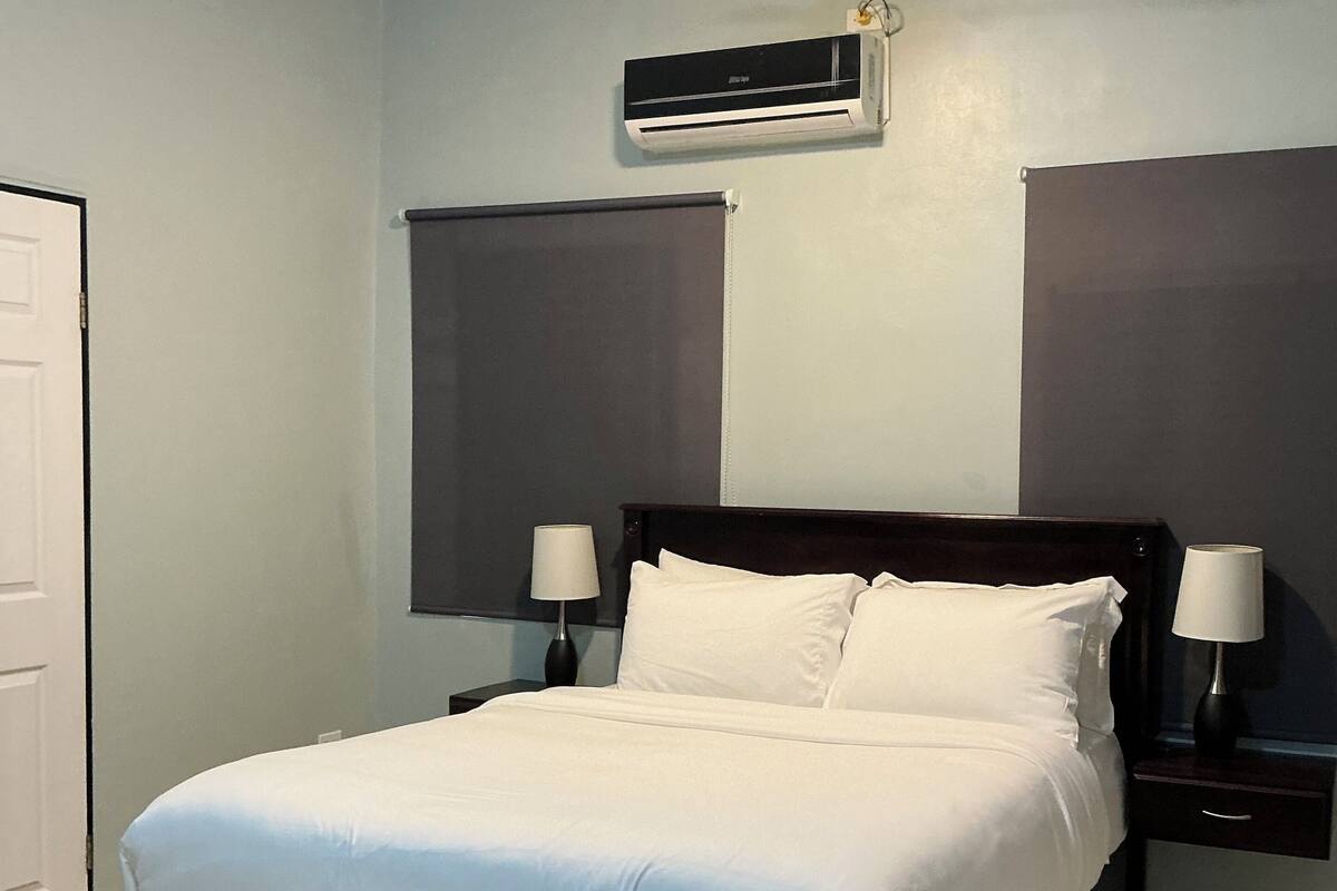 A bedroom with a bed, a ceiling fan, an air conditioning unit, two bedside tables with lamps, and a door.