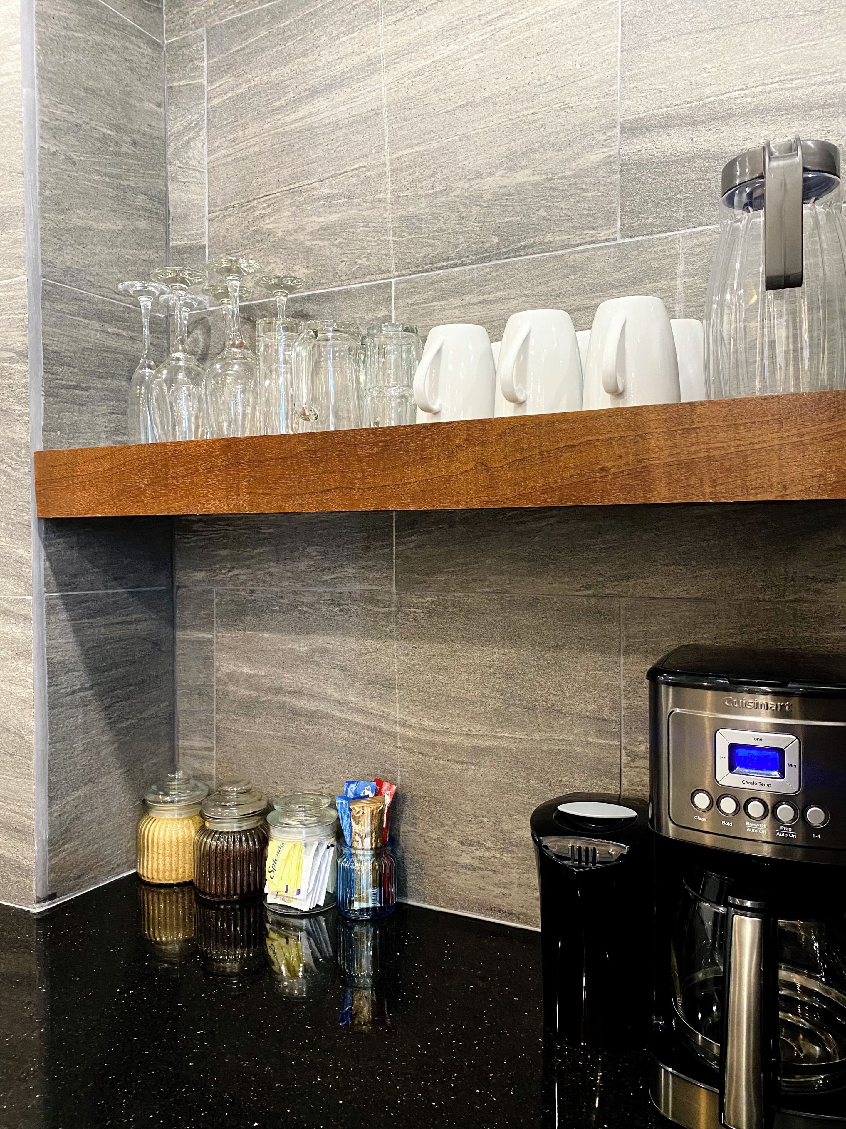 Un área de cocina con cafetera, repisas con vasos y tazas, y frascos de especias.
