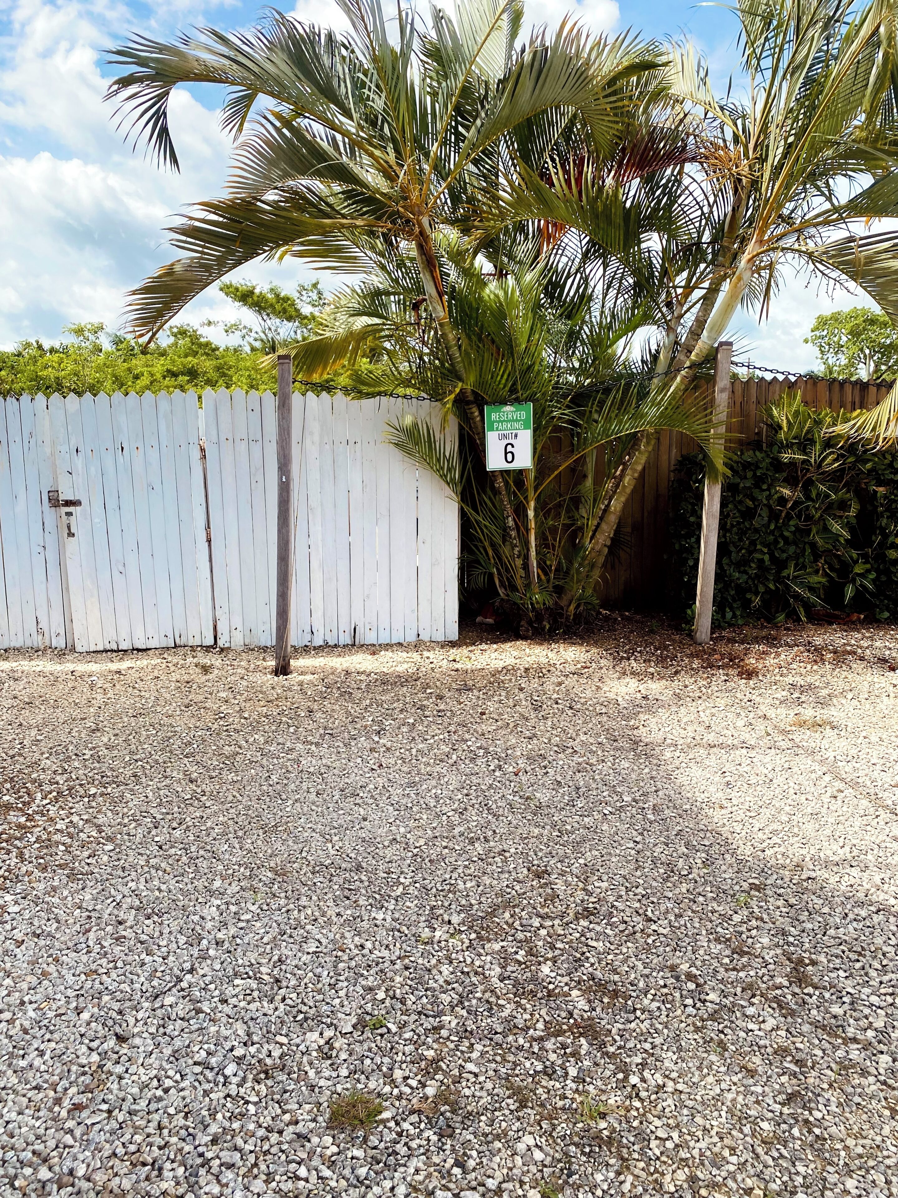 A gravel parking area with a white fence, a palm tree, and a sign indicating reserved parking unit #6.