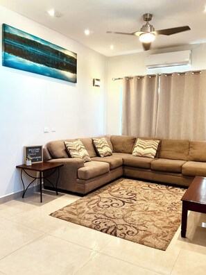 A living room with a sectional sofa, a patterned rug, a ceiling fan, and an air conditioning unit. - The Ansen Place (Belmopan)