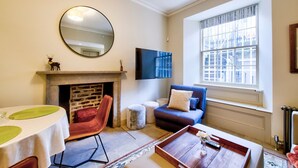 A living room with a fireplace, a round mirror, a television, a sofa, and a window with a view of buildings. - GuestReady - Manor Place Basement (Edinburgh)