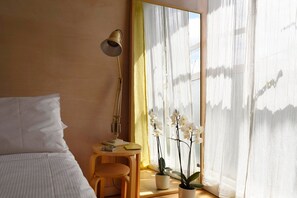 Une chambre à coucher avec un lit, un bureau avec une lampe, une chaise et une fenêtre avec des rideaux et des orchidées.