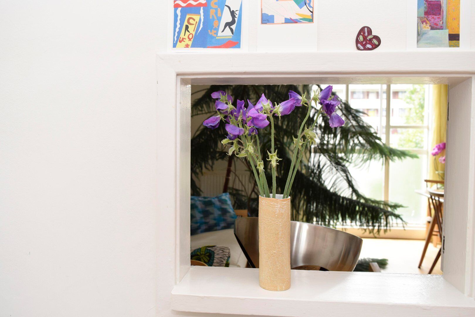 A window with a vase of purple flowers, a metal bowl, and a plant in the background.