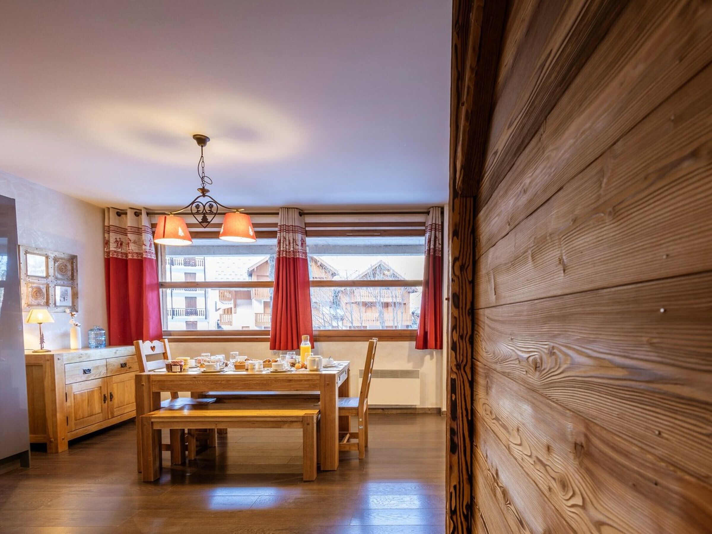 Una mesa de comedor de madera con sillas, una lámpara colgante y vistas de edificios a través de grandes ventanales.