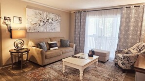 Ein Wohnzimmer mit einem beigefarbenen Sofa, einem weißen Couchtisch, gemusterten Vorhängen und einem großen abstrakten Gemälde an der Wand.