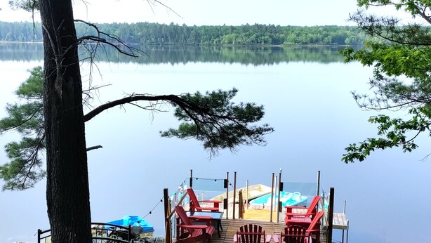 A dock with chairs and a small boat, overlooking a calm lake with trees in the background.